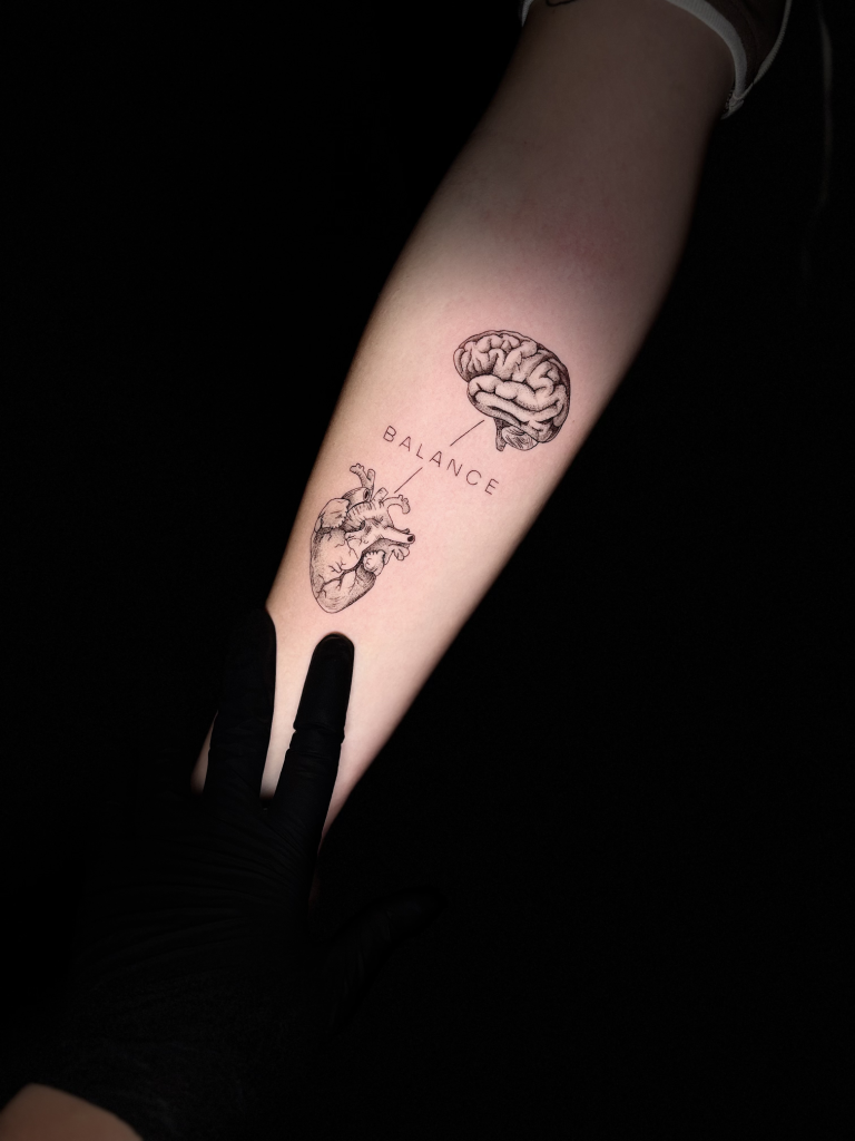 Tatuaje de línea fina en el antebrazo: un cerebro y una flor unidos bajo la palabra «balance»