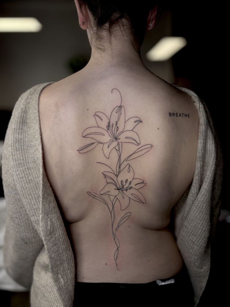 Lirio de línea fina de gran formato tatuado en la espalda, junto a la palabra «breathe»