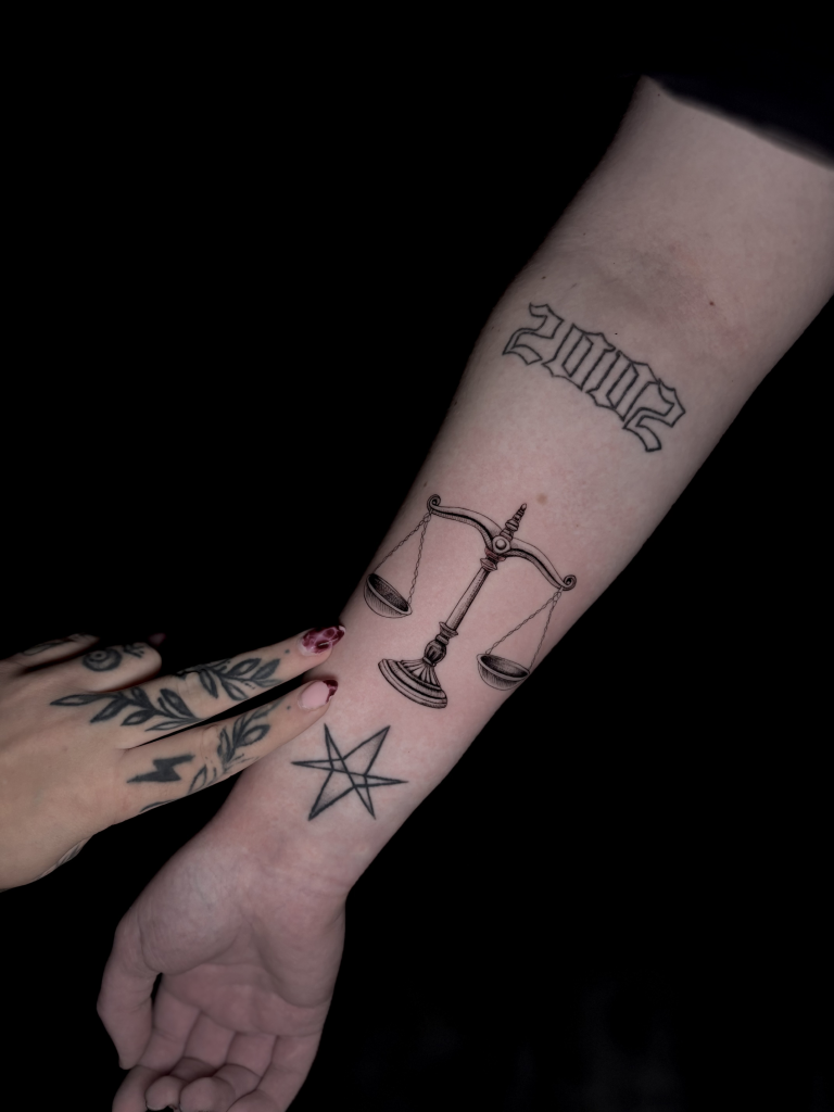 Antebrazo con varios tatuajes pequeños: la balanza de Libra, una estrella y la fecha 2002